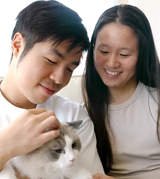 A young man and woman smile while petting a fluffy gray and white cat.