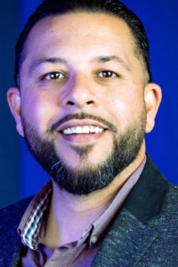 Man with dark hair and beard smiling with blue background