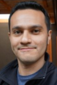 A man with short black hair smiles in a casual setting.