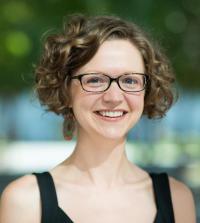 wearing glasses and with curly hair, Kassandra Ori-McKenney smiles and stares at the camera in an outdoor setting.