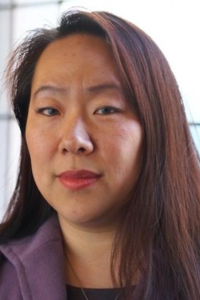 Woman with long dark hair and a serious expression, wearing a purple jacket.