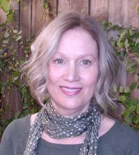 a woman with short blonde hair is wearing a grey patterned scarf and a grey blouse, in front of a wooden wall and floral background, staring at the camera