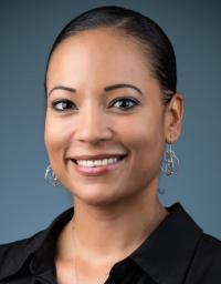 inside a photo studio, a woman smiles and stares at the camera for her profile photo. She's wearing a black top and has her hair back in a formal photo.