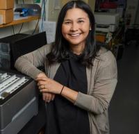 Megan Dennis smiles and stares for the camera within their laboratory. She is wearing a black shirt and grey cardigan, with long, dark hair parted at the middle touching her shoulders.