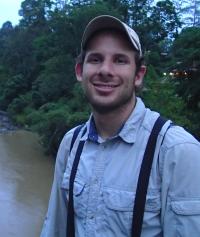outside near a small body of water at dusk, Daniel Karp is wearing a hat and facing the camera, smiling for a photo. He's wearing a plaid Oxford shirt and a baseball cap.