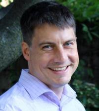 Smiling man in a light purple shirt, with greenery in the background.
