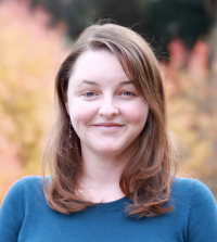 a woman with short brown hair and a teal sweater stares and smiles at the camera in an outdoor fall setting