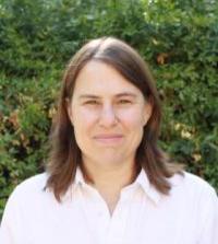 in an outdoor setting a woman with dark hair parted down the middle and a white Oxford shirt stares straight at the camera for a professional headshot