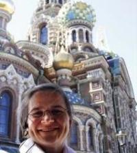 in front of a tall historic building stands a woman with pulled back hair and glasses smiling at the camera