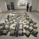 Floor installation of miniature houses on patches of green lawn along suburban streets, all on top of a map depicting American Indian symbols.