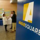 "Aggie Square" logo sign in foreground of meeting.