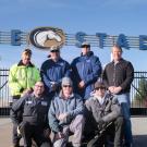 Seven men pose in front of Aggie Stadium gate and sign.