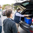 Two people watch as another psreon unloads blue box from vehicle.