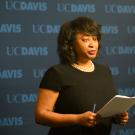 Woman reads from script against "UC Davis" backdrop.
