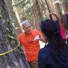 A man beside a tree lectures to onlooking fellows.