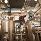 A man with a long beard and plaid shirt stands behind a row of silver metal tanks joined by pipes. 
