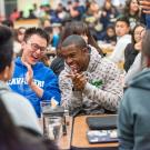 amidst a full indoor dining common, two students in the center frame of the photo are laughing next to each other. One is wearing glasses with a blue hoodie while another wears a grey top. 