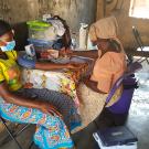 Two African women sit at a table. The women at left wears a blue patterned skirt, yellow top and black headscarf, and a blood pressure cuff on her arm. The woman at right wears brown clothes and a headscarf and is making notes on a form. 