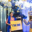 Student wears a blue feather boa poses with Gunrock during an Aggie Day celebration, holding a yellow “I’m In!” sign as blue and gold confetti falls.