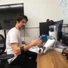 A young man in a white t-shirt sits at a desk and flexes his forearm, which has a band around it. On the desk is a robotic arm and a computer monitor. 