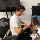 A person sits at a desk with a robotic arm, smiling and making a fist.
