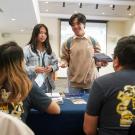 Two students, one in a tan sweater and the other in a jean jacket, stand in front of a table and ask questions to two UC Davis representatives in navy short sleeve Global Aggie shirts.  