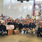 Eight people pose for photo in front of large pile of canned food and other supplies