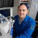 A man in a dark blue labcoat sits in front of a large microscope looking up towards the camera. 