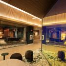 An angular view of two brightly lit, glass offices converging in a carpeted common space with chairs and a table.