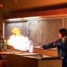 Inside a lecture hall, towards the right of the frame, a student in a lab coat with goggles on holds a stick that is engulfed in flames. The flames are caused by the demonstration the student is leading to a crowd at Rock Hall on UC Davis. On the left, a man in a lab coat plugs his ears at the sound of the controlled explosion. 