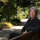 In an outdoor setting, Kate Scow sits on a bench towards the right of the photo, facing the camera and smiling. She's wearing a dark blue long sleeve top and is angled just slightly toward the camera. 