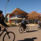 On a sunny fall day, three students on bicycles ride along a path with the restaurant Latitude in the background.