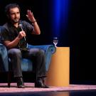 A speaker, Javier Zamora, gestures while seated on a blue chair, water glass nearby, under stage lights.
