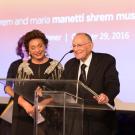 Woman and man in evening attire, at microphone, in front of backdrop of Manetti Shrem Museum sign in rainbow colors