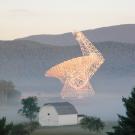 A large radiotelescope dish, white but lit by golden hour light, against a background of misty forested mountains. 