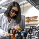 A woman with dark hair wearing a white lab coat and dark wraparound eyeglasses leans over technical equipment on a lab workbench. 