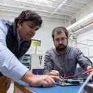 Two young men lean over a work table. Person on right, with short beard and glasses, is using a tool to test a circuit board as the person on left, with dark hair wearing a shirt and sleeveless vest, leans to examine it. 