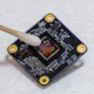 A black square with printed circuitry, tilted at an angle, against a white background. A wet q-tip points at the center of the square. 