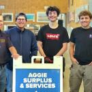 From left to right, AggieSurplus Coordinator Robert Juhler and Student Assistants Nick Gomez, Isaac Ewing and Lucas Morace stand and smile behind a sign that says "Aggie Surplus and Services." 