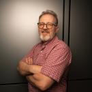 Professor Andreas Bäumler stands for a portrait with his arms crossed at a slight angle towards the camera, against a white backdrop inside an office setting. He is wearing a plaid red short sleeve shirt, glasses and has a full beard with hints of grey, and is smiling towards the camera. 