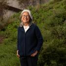 Outdoors in front of a grassy hill stands Professor Isabel Montañez, smiling towards the camera. She is wearing glasses, a blue button-up shirt with white undershirt and black pants. 