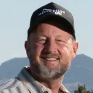A smiling man with a beard wearing a cap and a striped shirt, standing outdoors with mountains in the background.