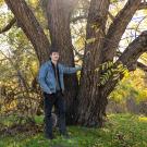 A man wearing a denim jacket and dark pants stands under a large tree with multiple spreading limbs. 