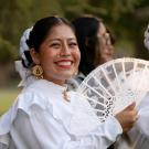 Two women in traditional white attire smile while holding lace fans in a sunny outdoor setting.