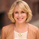 Smiling woman with blonde hair, wearing a beige jacket and gold necklace, outdoors.