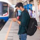 A man and woman wearing masks and looking at their phones wait to board an approaching train