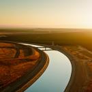 Drone shot of California aqueduct curving between an auburn barren hillside and agricultural fields in Kern County at sunset