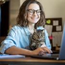 Woman using a laptop with a relaxes cat on her lap, representing how telehealth helps pet owers manage chronic conditions in cats from home. 
