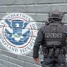 Photo of ICE agent in front of gray wall that says Department of Homeland Security via a logo