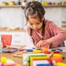 A young girl with braids wearing a pink top concentrates on playing with blocks. 